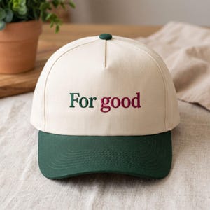 May include: A cream-coloured baseball cap with a dark green brim and a dark green button on top. The front of the cap features the embroidered text "For good" in a serif font, with the word "For" in dark green and "good" in a deep maroon colour.