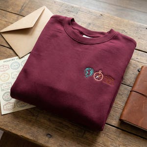 May include: A maroon sweatshirt with embroidered travel-themed icons: a globe, compass, and camera. The sweatshirt is folded on a wooden surface, with a passport stamp sheet, envelope, and leather journal nearby.