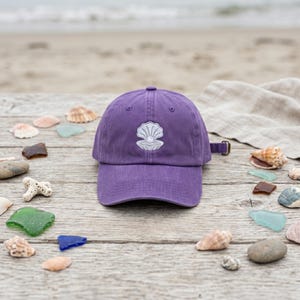 May include: A purple baseball cap with a white embroidered seashell design. The cap is made of a soft material and has an adjustable strap. The background includes seashells, sea glass, and a beach scene.