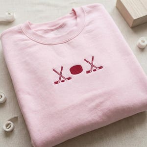 May include: A light pink sweatshirt with a crew neck. The front of the sweatshirt features an embroidered design of two crossed hockey sticks and a hockey puck in red. The design is repeated on the right side.