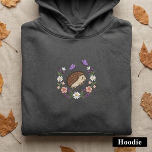 May include: Dark gray hoodie featuring an embroidered design of a hedgehog surrounded by a floral wreath with pink flowers, white daisies, and purple butterflies. The word "Hoodie" is printed at the bottom.