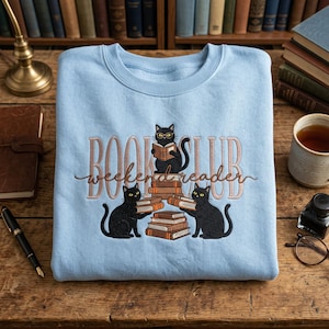 May include: A pale blue sweatshirt with the embroidered text "BOOK CLUB weekend reader." Three black cats are depicted, one reading a book on a stack of books, and two holding books. The sweatshirt is on a wooden surface.