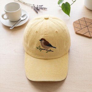 Peut inclure: Une casquette de baseball jaune pâle ornée d'une broderie représentant un moineau brun perché sur une fine branche verte. La casquette présente une calotte structurée avec un bouton au sommet, des œillets cousus et une visière courbée.