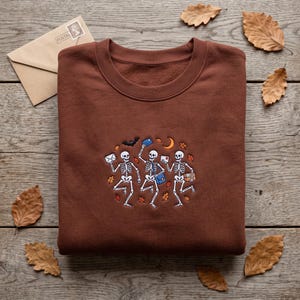 May include: A brown crewneck sweatshirt featuring an embroidered graphic of three white skeletons carrying mail, a blue satchel, and a package. The design includes a black bat, a yellow crescent moon, and scattered orange and brown autumn leaves.