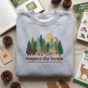 Könnte beinhalten: Ein hellgraues Sweatshirt mit einer gestickten Waldszene. Es zeigt einen Bären, einen Fuchs, einen Waschbären, ein Reh, eine Eule und ein Kaninchen. Der Text "respect the locals" und "explore and protect their natural habitat".