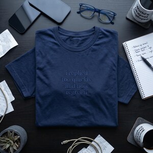 May include: Navy blue t-shirt with the embroidered text "i replied too quickly and now regret it." The shirt is displayed on a dark surface with office supplies, including glasses, a notepad, and a coffee mug.