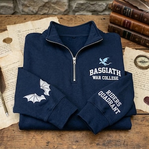 May include: A navy blue quarter-zip sweatshirt. It features white embroidered bat wings on the left sleeve and the words "BASGIATH WAR COLLEGE" and "RIDERS QUADRANT" in white. The sweatshirt has a silver zip.
