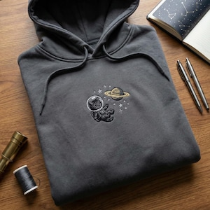 May include: Charcoal grey hoodie with a drawstring hood. The front displays an embroidered design of a cat in an astronaut suit floating near a planet with rings and stars. The hoodie is on a wooden surface.