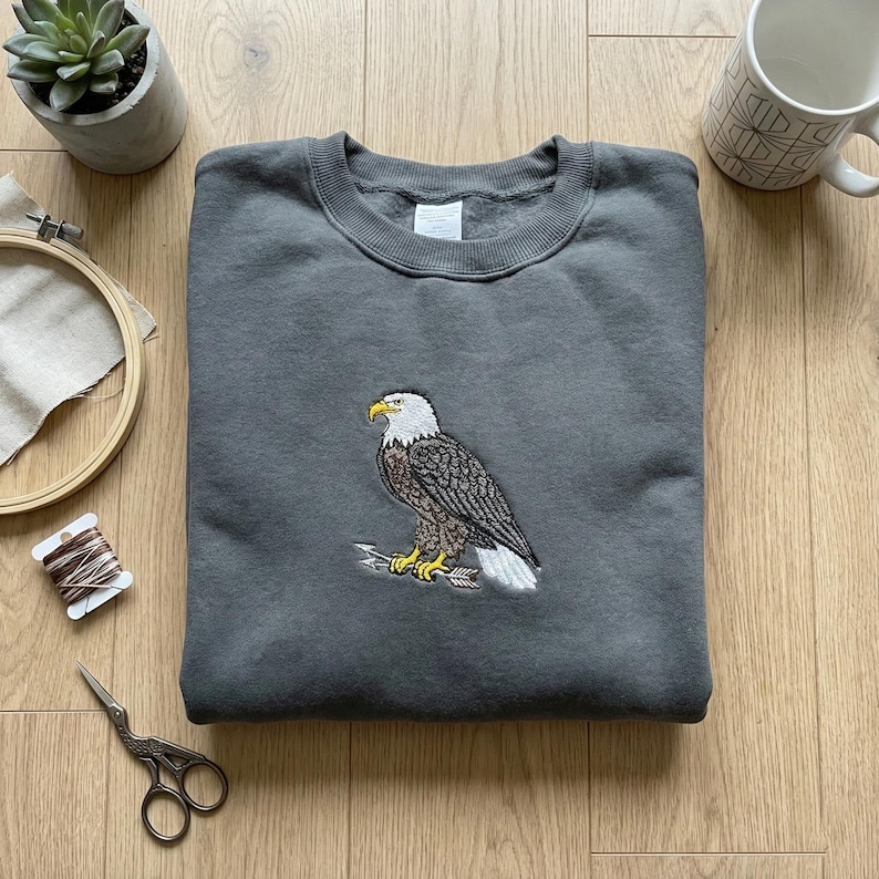 May include: A charcoal grey sweatshirt featuring an embroidered bald eagle. The eagle design includes white, brown, yellow, and black details, perched on arrows. The sweatshirt is neatly folded on a wooden surface.