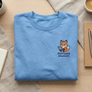 May include: A light blue sweatshirt with a small embroidered design of a fox using a laptop. The text "SOFTWARE ENGINEER" is below the fox. The sweatshirt is folded neatly on a light-colored surface.