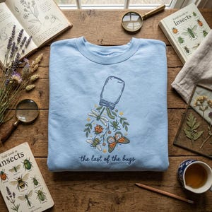 Könnte beinhalten: Ein hellblaues Sweatshirt mit Rundhalsausschnitt und einer Stickerei, die ein Glas zeigt, aus dem verschiedene Insekten wie ein Schmetterling, ein Marienkäfer, eine Libelle und eine Biene herauskommen, mit dem Schriftzug "the last of the bugs".