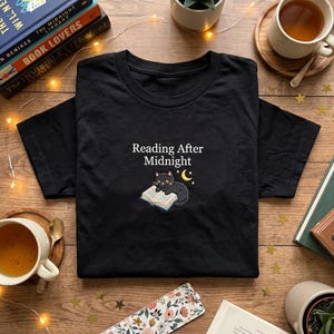 May include: Black t-shirt with the embroidered text "Reading After Midnight" and a cartoon cat reading a book under a crescent moon and stars. The shirt is displayed on a wooden surface with books, tea, and decorative items.