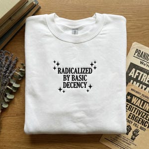 May include: A white crew neck sweatshirt featuring the embroidered phrase "RADICALIZED BY BASIC DECENCY" in black, with starburst accents. The garment is neatly folded, resting on a wooden surface.