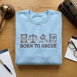 Op de afbeelding: Een lichtblauw sweatshirt met de geborduurde tekst "BORN TO ARGUE" onder juridische symbolen. Deze omvatten een zandloper, weegschaal, hamer en een boek met het woord "LAW".