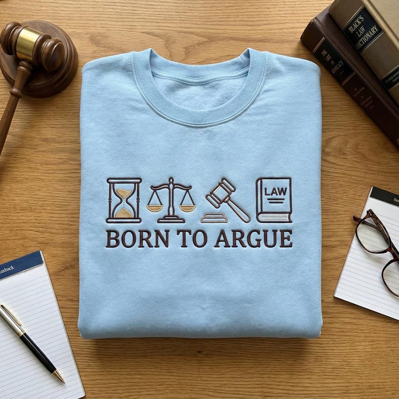 Op de afbeelding: Een lichtblauw sweatshirt met de geborduurde tekst "BORN TO ARGUE" onder juridische symbolen. Deze omvatten een zandloper, weegschaal, hamer en een boek met het woord "LAW".