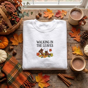 May include: A white crewneck sweatshirt featuring a central graphic with the text WALKING IN THE LEAVES in black, accompanied by an illustration of brown boots surrounded by colorful autumn leaves in shades of orange, yellow, and green.