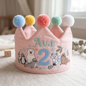 May include: A light pink fabric crown featuring the embroidered name Ava and the number 2. The design includes arctic animal illustrations of a penguin, polar bear, seal, fox, and walrus, accented with snowflakes and topped with five colourful pom-poms.