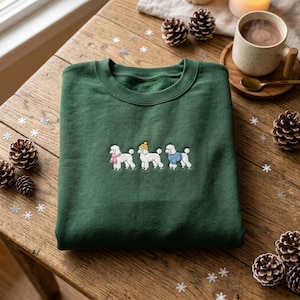 May include: A dark green crewneck sweatshirt featuring an embroidered design of three white poodles. The left poodle wears a pink scarf, the centre poodle wears a yellow beanie, and the right poodle wears a blue jumper. The fabric is solid green.