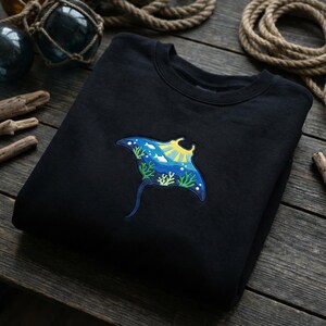 Peut inclure: Sweat-shirt noir orné d'une broderie de raie manta colorée. La raie manta est bleue avec un soleil jaune, des poissons blancs et des coraux verts. Le sweat-shirt est plié sur une surface en bois.