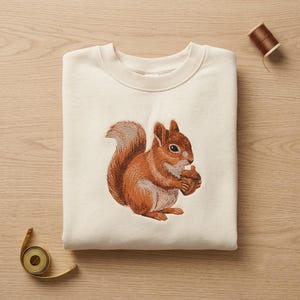 May include: A cream-coloured crewneck sweatshirt featuring a detailed embroidered graphic of a brown squirrel holding an acorn. The design is centred on the chest. A small spool of brown thread and a yellow measuring tape are visible nearby.