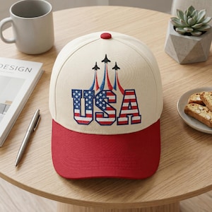 May include: A cream-colored baseball cap with a red brim and a red button on top. The front features the text USA in a red, white, and blue flag pattern, with three black jet silhouettes above it trailing red and blue smoke plumes.