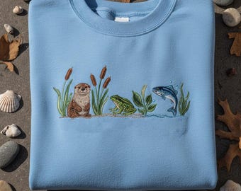 Op de afbeelding: Lichtblauw sweatshirt met een geborduurd ontwerp van een otter, kikker en vis in een wetland-omgeving. Het ontwerp bevat lisdodden en waterplanten.