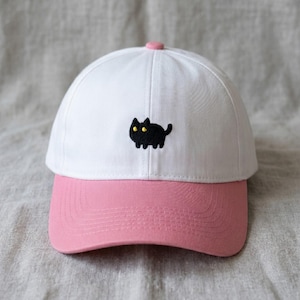 May include: White baseball cap with a pink peak and button. A black cat with yellow eyes is embroidered on the front. The cap is made of a soft fabric and is suitable for casual wear.