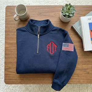 May include: Navy blue quarter-zip sweatshirt with a red embroidered monogram and an American flag patch on the sleeve. The sweatshirt has a high collar and a silver zipper. A casual, comfortable top.