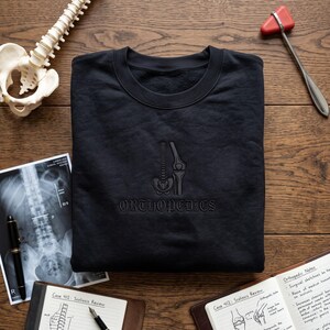 May include: A black crewneck sweatshirt featuring an embroidered graphic of a human spine, pelvis, and knee joint, with the word ORTHOPEDICS stitched in a dark thread below the anatomical illustration, centred on the chest of the garment.