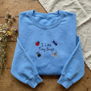 May include: A light blue sweatshirt with the embroidered text "I Like Tiny Bugs" and illustrations of a ladybug, ant, beetle, and moth. The crew neck sweatshirt is made of a soft material.