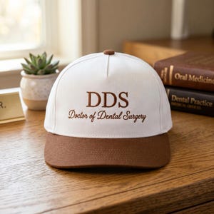 May include: A white and brown baseball cap with the text "DDS Doctor of Dental Surgery" embroidered in brown. The cap has a brown brim and button, and is sitting on a wooden surface.