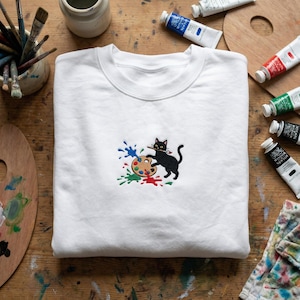 May include: White crewneck sweatshirt featuring an embroidered black cat holding a paintbrush and a colorful artist's palette. Splashes of blue, green, and red paint surround the cat and palette. The sweatshirt is on a wooden surface with art supplies.