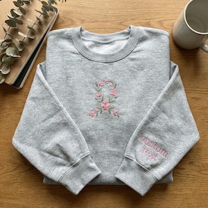 May include: A grey sweatshirt featuring a pink and green floral embroidery on the chest. The right sleeve displays the words "custom text" in pink. The garment is folded on a wooden surface.