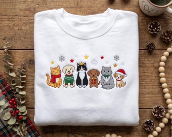 May include: A white crewneck sweatshirt featuring an embroidered design of six cartoon animals including cats and dogs wearing holiday accessories like a red scarf, green sweater, gold crown, and Santa hat, accented by embroidered stars and snowflakes.