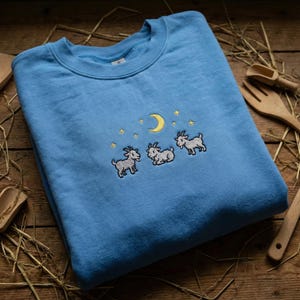 May include: A light blue sweatshirt featuring an embroidered design of three goats, a crescent moon, and stars. The crew neck sweatshirt is made of a soft material and is displayed on a wooden surface with hay and wooden kitchen utensils.