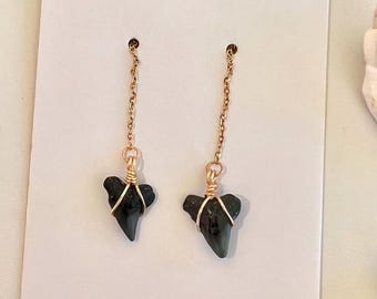 May include: A pair of drop earrings featuring dark gray shark teeth pendants wrapped in thin gold-toned wire. Each tooth is suspended from a delicate gold-toned metal chain, presented on a plain white rectangular display card with a top hole.