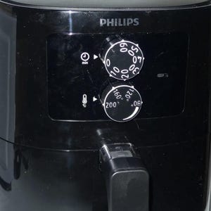 May include: A black Philips air fryer featuring a glossy finish, a front-facing handle, and a control panel with two circular dials. The top dial controls time in minutes, while the bottom dial adjusts temperature in degrees Celsius from 80 to 200.