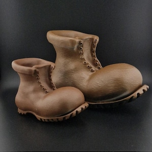 May include: Two brown ceramic boots with laces and a textured surface. The boots are different sizes, with the larger boot in the background and the smaller boot in the foreground.