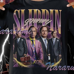 Peut inclure: T-shirt noir avec un graphisme rétro de quatre hommes en costume et le texte "SLIPPIN' JIMMY". Le design utilise une palette de couleurs violet et or avec des éclairs.