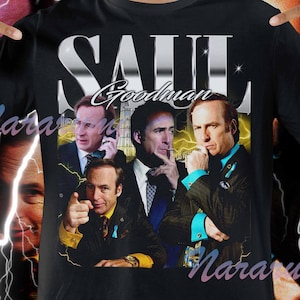 Peut inclure: T-shirt noir avec le nom "SAUL Goodman" en lettres argentées et un collage d'images. Le collage présente un homme en costume. Le t-shirt a un col rond et des manches courtes.