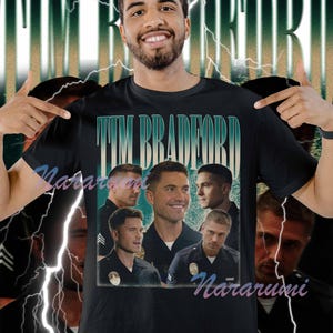 May include: Black t-shirt featuring a collage of images and the text "TIM BRADFORD" in teal and white. The shirt has a vintage aesthetic with a faded background and lightning bolt graphics.