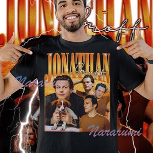 Puede incluir: Camiseta negra con un gráfico retro de Jonathan Groff, con múltiples imágenes de él. El diseño incluye el nombre "Jonathan Groff" en una fuente dorada vintage. La camiseta tiene cuello redondo y mangas cortas.