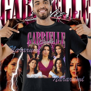 Op de afbeelding: Zwart t-shirt met een retro ontwerp in roze en wit met de naam "GABRIELLE Solis". Het shirt toont een afbeelding van Gabrielle Solis, wat een vintage esthetiek creëert.