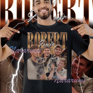 May include: Black t-shirt featuring the name "ROBERT IRWIN" in gold lettering. The shirt includes a collage of images of Robert Irwin holding a koala and smiling. The background has a vintage design with lightning bolts.