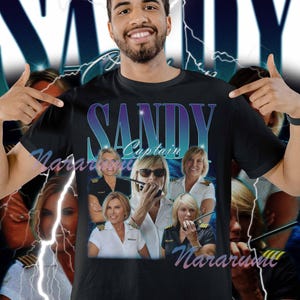 May include: Black t-shirt featuring a graphic of Captain Sandy with the text "SANDY Captain" in a gradient blue and purple font. The shirt has a collage of images of Captain Sandy.