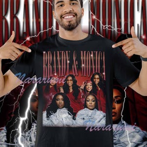 Pode incluir: Camiseta preta com um gráfico de Brandy e Monica. O design inclui uma foto em grupo das cantoras em vermelho e branco, com o texto "Brandy & Monica" em uma fonte vermelha vintage. O fundo tem um design vermelho e branco com raios.