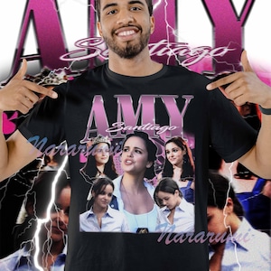 以下が含まれることがあります： 黒いTシャツ。ピンク色の「AMY Santiago」の文字と女性の複数の画像が特徴です。デザインには、背景にピンク色の稲妻模様と「Santiago」の文字も含まれています。