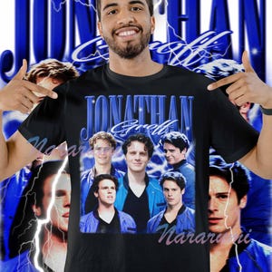 Puede incluir: Camiseta negra con un diseño azul que presenta el nombre "Jonathan Groff" y un collage de imágenes. El diseño incluye un rayo azul y la palabra "Narari" en rosa. La camiseta la lleva una persona.