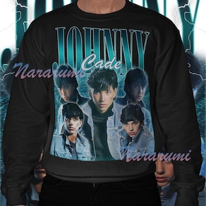 May include: Black sweatshirt featuring a teal and white graphic with the text "JOHNNY Cade" and images of a young man. The design has a vintage, retro aesthetic. The sweatshirt is a crew neck style.