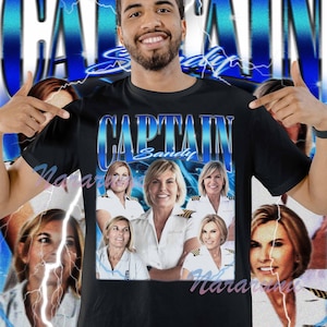Op de afbeelding: Zwart t-shirt met de tekst "CAPTAIN Sandy" in een blauwe gradiëntkleur. Het shirt bevat afbeeldingen van een vrouw in een kapiteunsuniform. De achtergrond heeft een blauw bliksemontwerp.